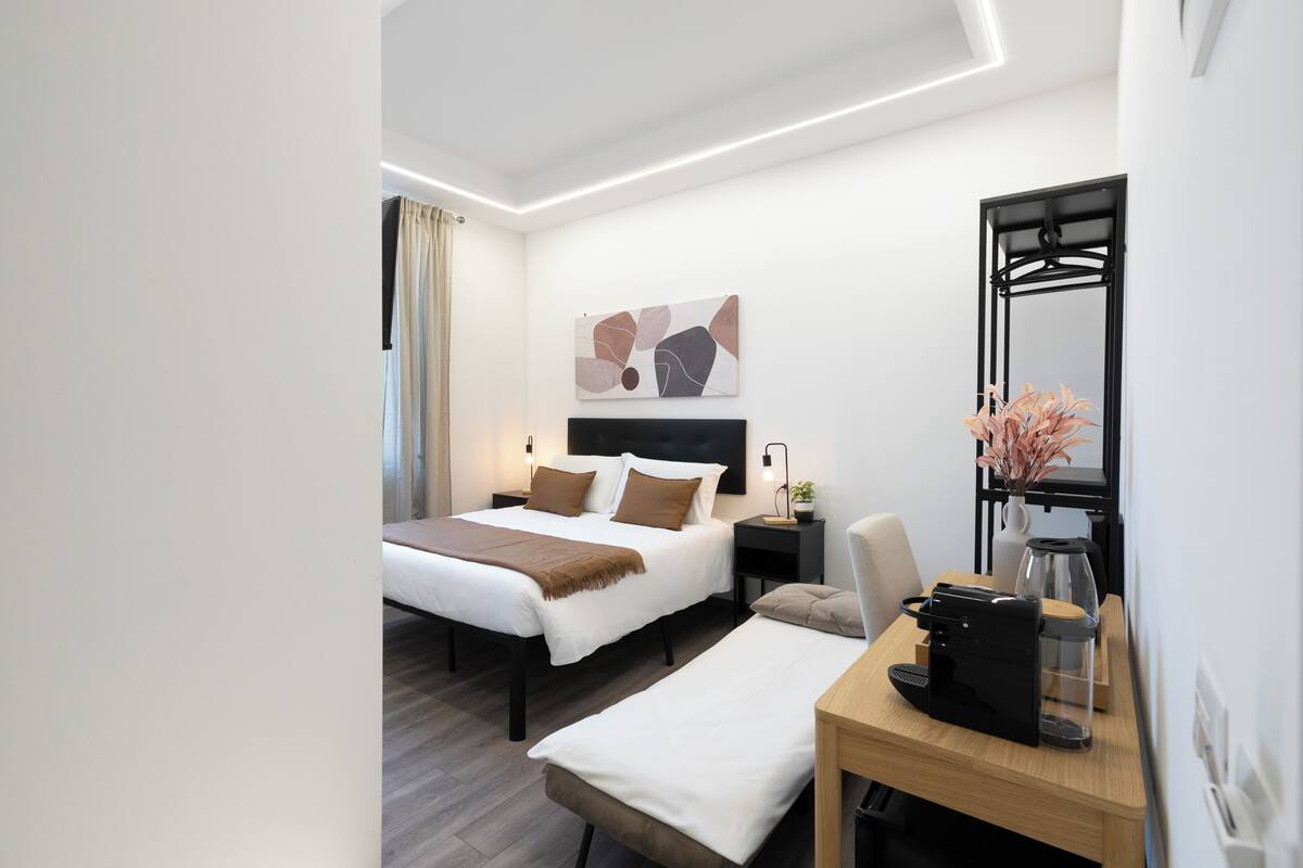 A modern hotel room with a bed, bedside tables, a desk, a mirror, and a wall art piece.