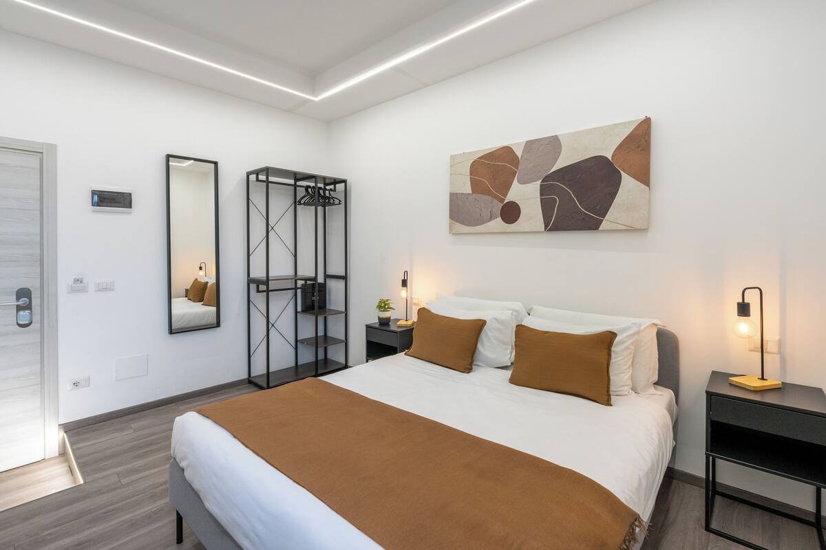 A modern hotel room with a large bed, a wardrobe, a bedside table with a lamp, and a wall-mounted abstract painting.
