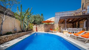 Una piscina con sillones de descanso y una sombrilla, rodeada de muros de piedra y cactus.