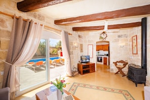 A stone-floored living room with a TV, wooden furniture, a fireplace, and a view of a swimming pool.