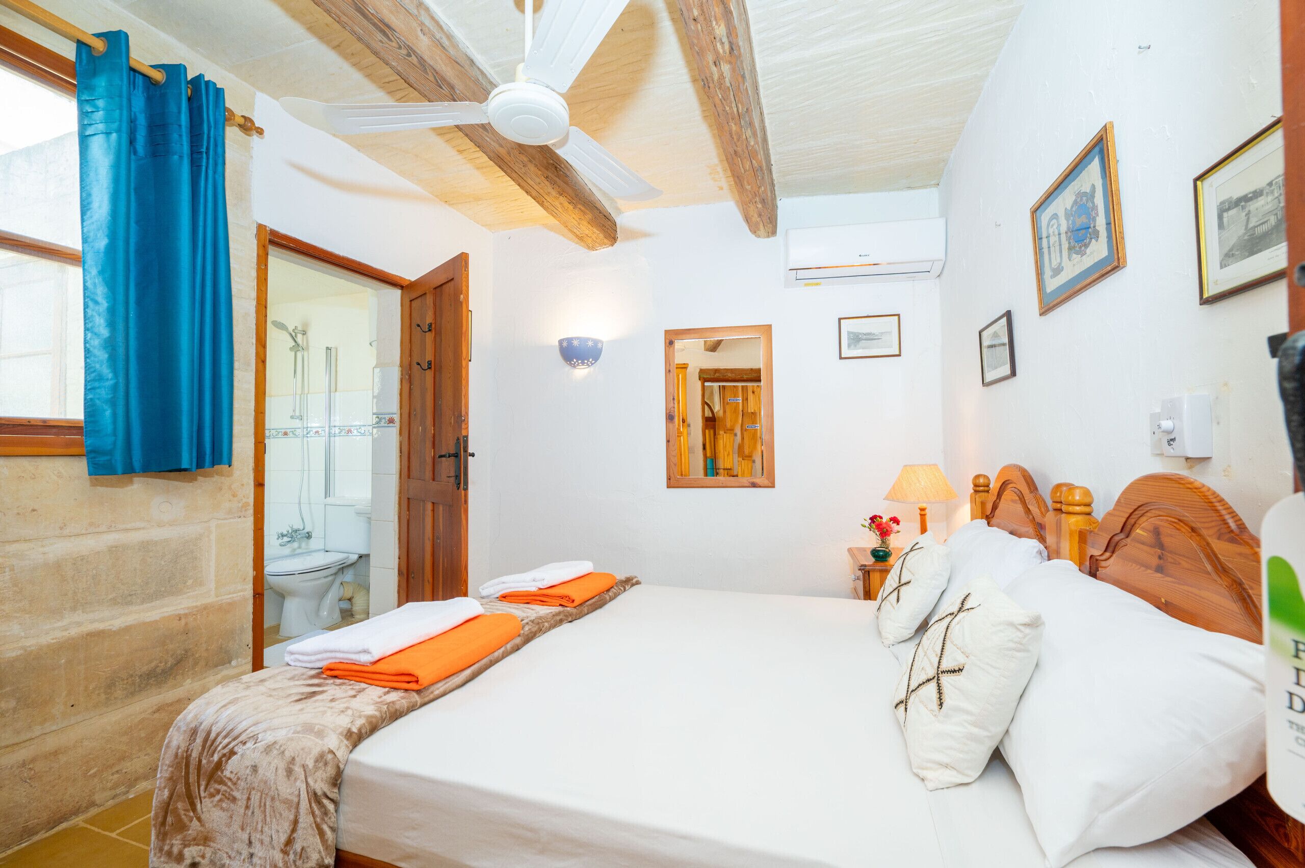 Une chambre à coucher avec un lit, un ventilateur de plafond, une tête de lit en bois, une fenêtre avec des rideaux bleus et une salle de bain visible à travers une porte ouverte.