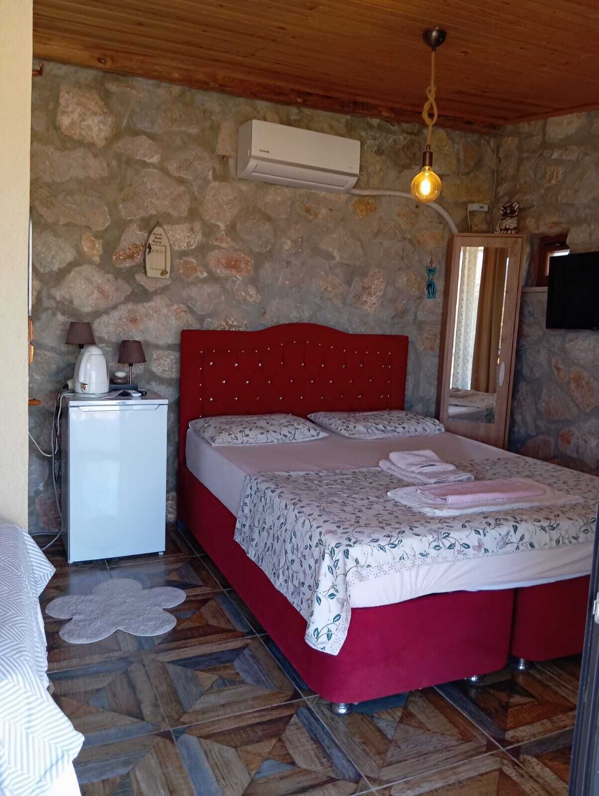 Un dormitorio con pared de piedra, una cama con cabecera roja, una mesita de noche blanca con lámparas, un refrigerador pequeño y una lámpara colgante.