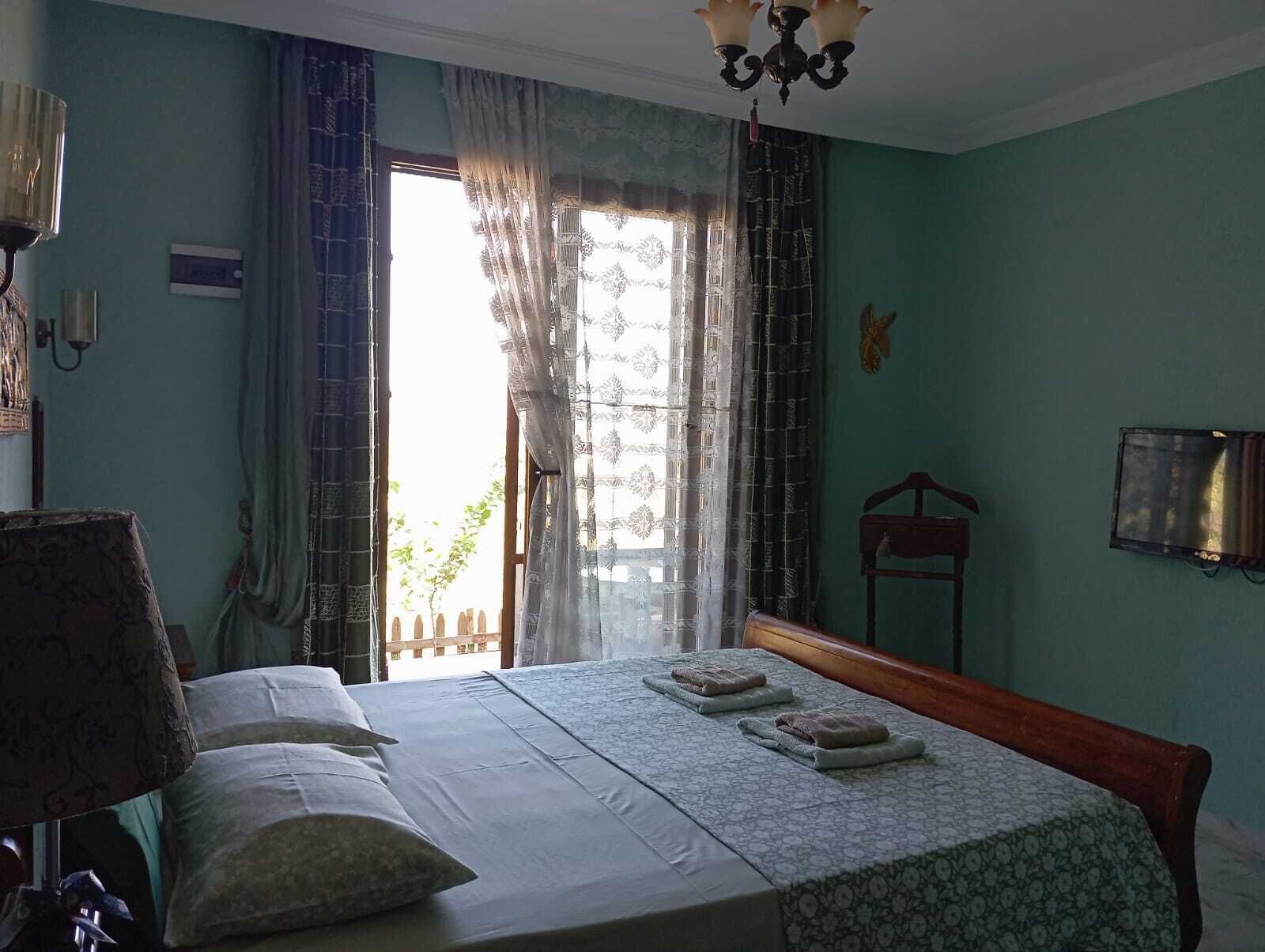 Un dormitorio con cama, una silla, un televisor y una ventana con cortinas.