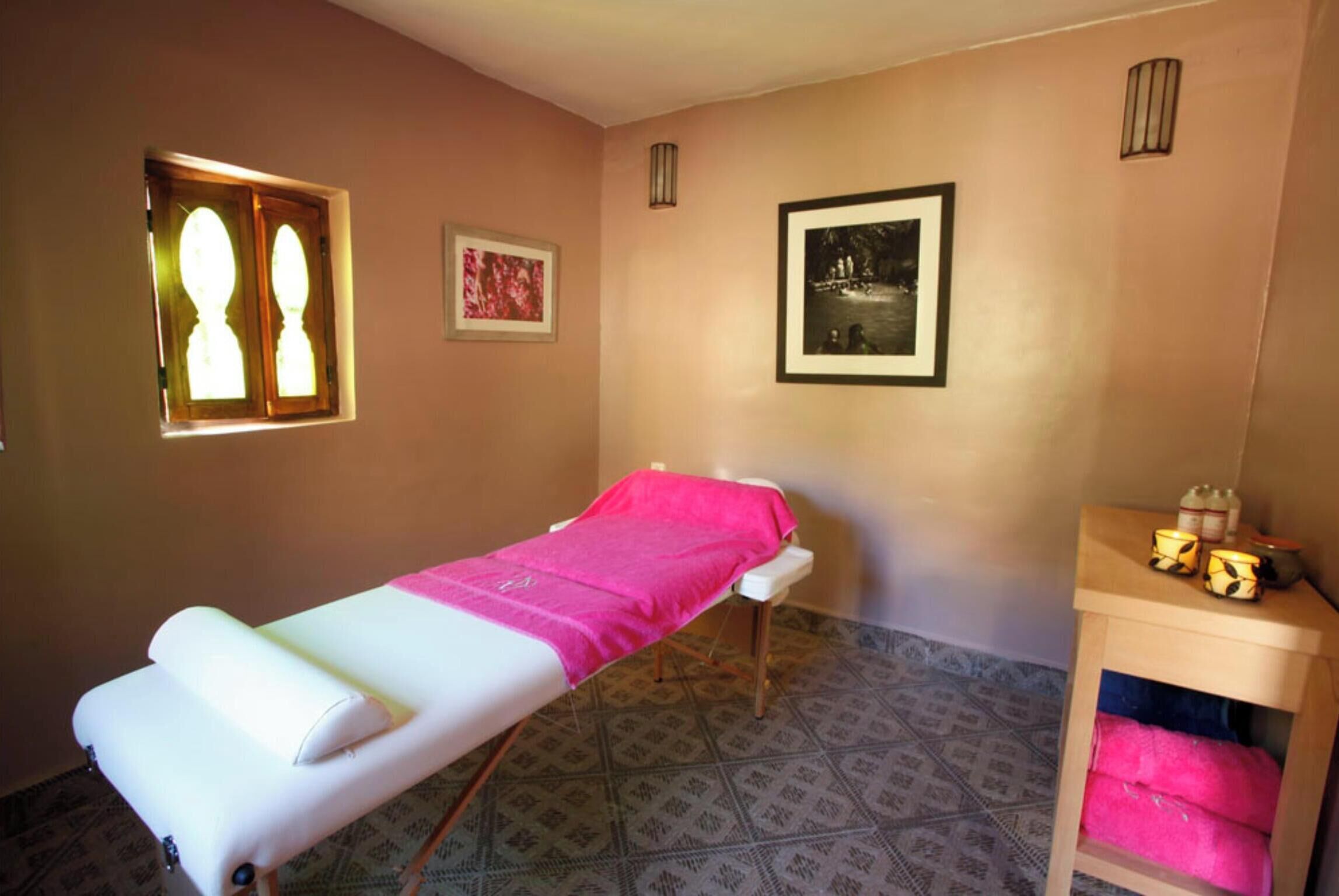 A room with a single bed, a small table with a pink towel, a framed picture on the wall, and a window with a view.