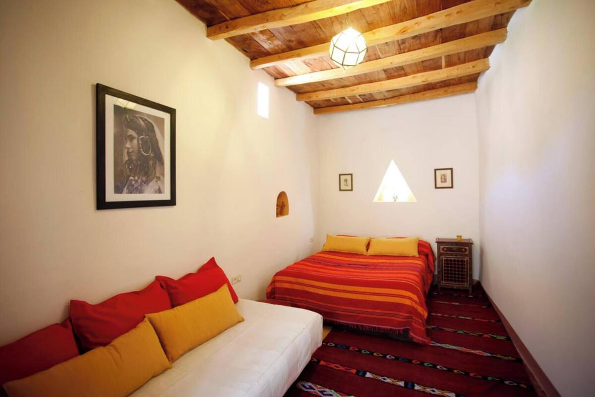 Ein Zimmer mit Holzbalkendecke, zwei Betten mit roter und gelber Bettwäsche, einem weißen Sofa und einem gerahmten Bild an der Wand.