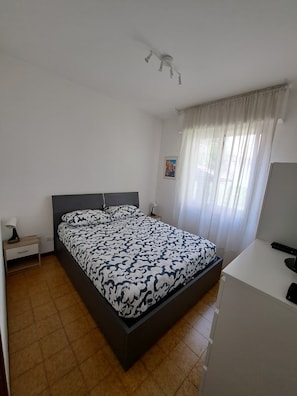 A bedroom with a bed, a bedside table, a desk, and a window with curtains. - USCIO NATURA DESCRIPTION (Uscio)