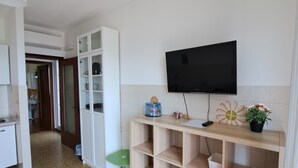 Ein Zimmer mit an der Wand montiertem Fernseher, einem Holzkabinett mit Schubladen, einem grünen Hocker und einem Fliesenboden.