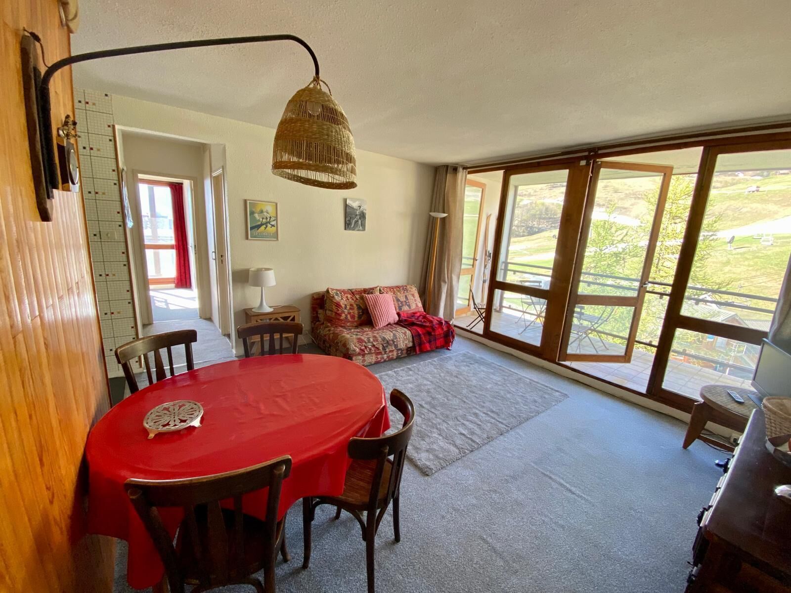 Un comedor con una mesa y sillas rojas, una lámpara de mimbre, un sofá con una manta estampada y un ventanal con vistas a un paisaje verde.