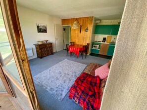 Ein Zimmer mit Esstisch, Stühlen, einer roten Tischdecke, einer Küchenzeile mit einem grünen Schrank und einer gemusterten Decke auf dem Boden.