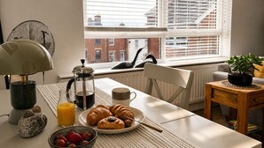 En frukostuppsättning med croissanter, en kaffekanna och en skål med frukt på ett bord vid ett fönster med persienner.