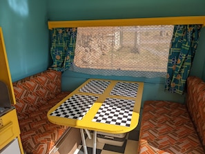 Interior d'una caravana compacta amb taula estampada, seients amb coixins, finestra amb cortines decoratives i un marc de finestra groc.