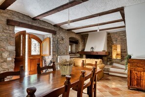 Zona de menjador rústica amb taula i cadires de fusta, parets de pedra i una llar de foc.