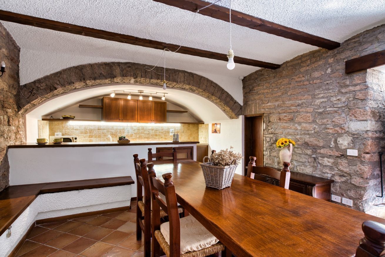 A rustic dining area with a wooden table and chairs, a stone wall, and a kitchen with wooden cabinets and a tiled backsplash.