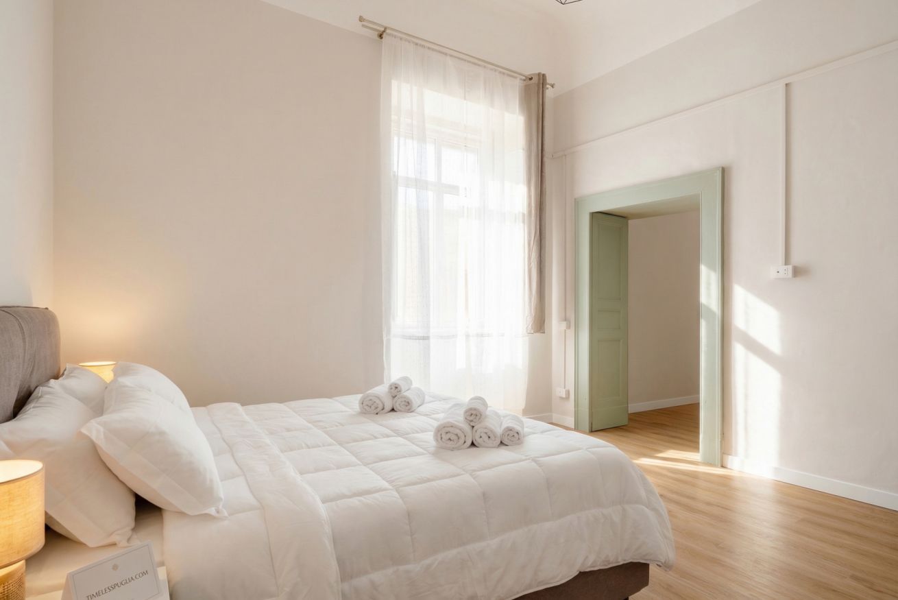 Un dormitorio con cama, almohadas, una mesita de noche, una lámpara y una ventana con cortinas.