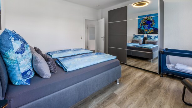 Een moderne slaapkamer met een grijze slaapbank, een blauw kussen met patroon, een grote spiegel en een blauwe fauteuil.