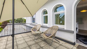 En terrasse med to fletstole og et bord, en beige parasol samt en bygning med buede vinduer.