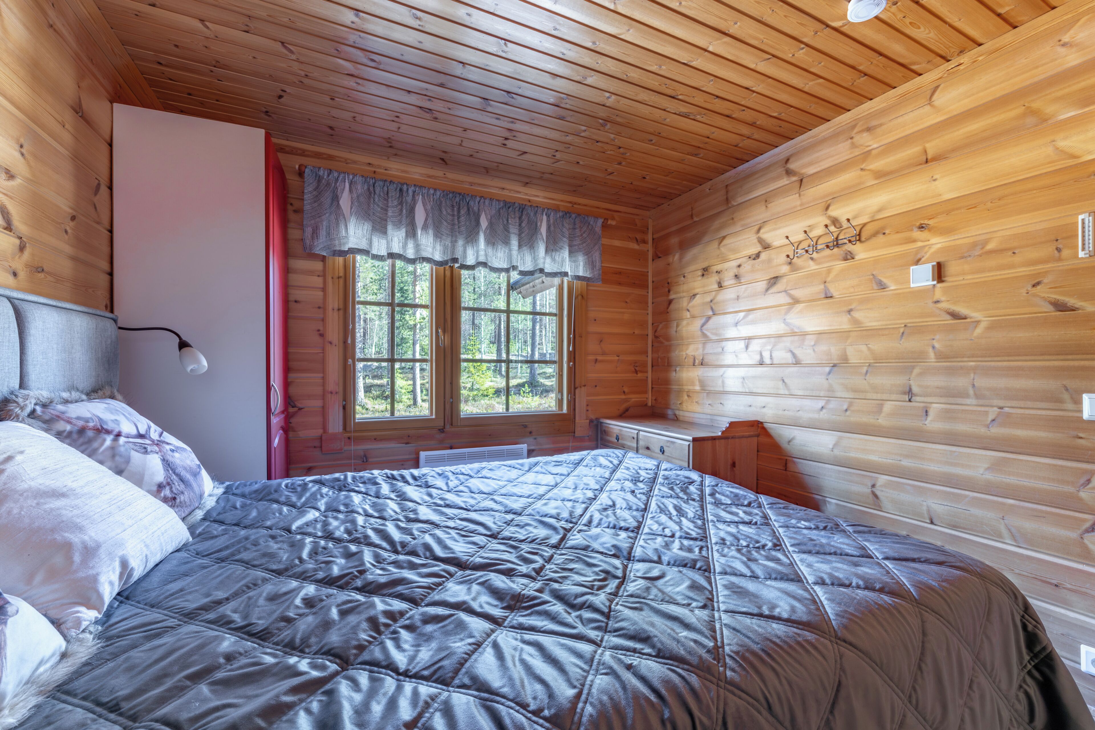 A wooden cabin bedroom with a bed, a window with a view of trees, a dresser, and a wall-mounted light fixture.