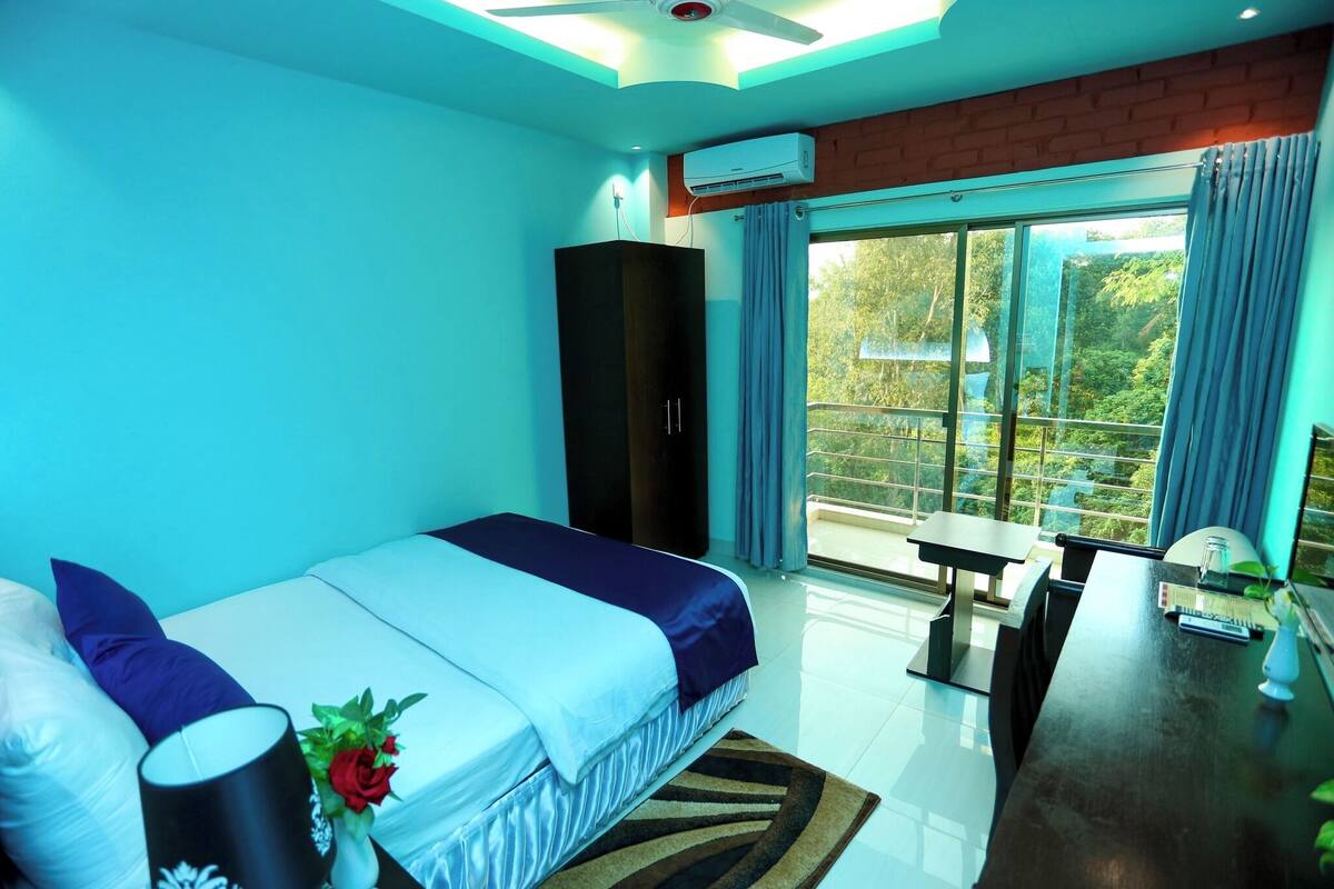 Un dormitorio con cama, escritorio, armario y una ventana grande con vistas a zonas verdes.