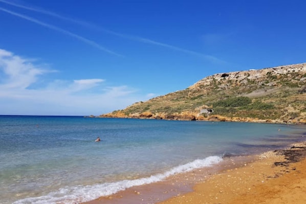 A beach with clear water, a sandy shore, and a rocky hill in the background. - Charming Gozo Escape | Balcony • AC • Fast Wi-Fi (Iż-Żebbuġ)