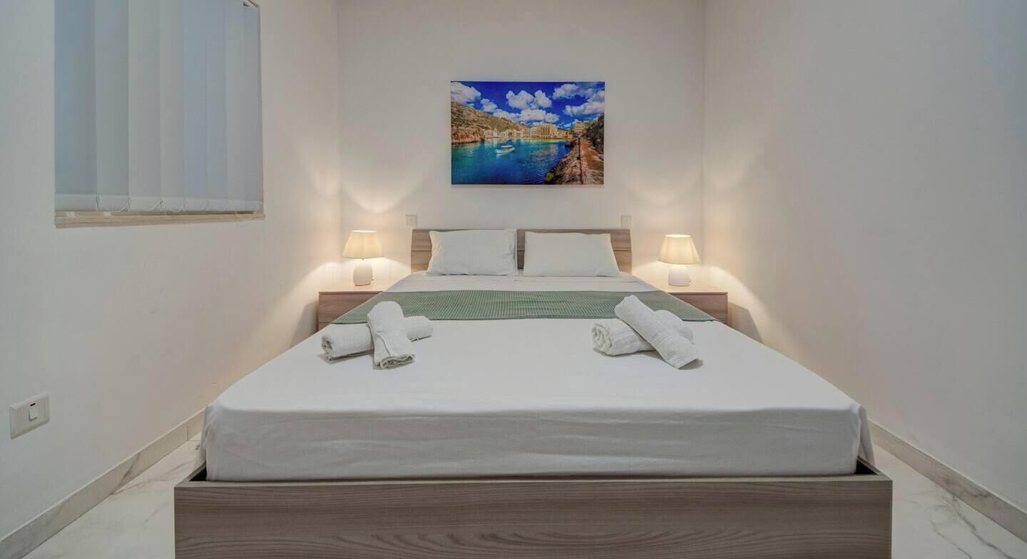 A neatly made bed with white linens and two rolled towels, a painting of a coastal scene above it, and two bedside lamps on either side.