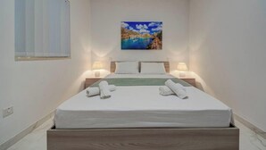Un letto rifatto con lenzuola bianche e due asciugamani arrotolati, un quadro raffigurante una costa sopra il letto, e due abat-jour ai lati.