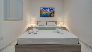A neatly made bed with white linens and two rolled towels, a painting of a coastal scene above it, and two bedside lamps on either side.