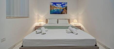 A neatly made bed with white linens and two rolled towels, a painting of a coastal scene above it, and two bedside lamps on either side.