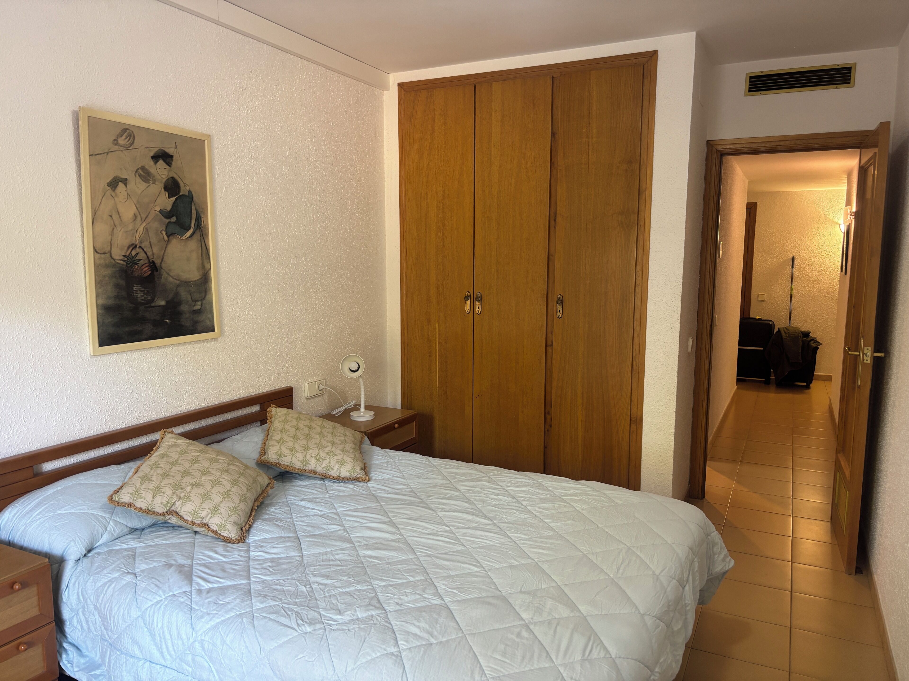 A bedroom with a bed, a nightstand, a wooden wardrobe, a painting, and a hallway.