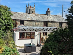 A stone house with a small attached structure featuring a glass door and windows, a tiled roof, and a garden area with a table and potted plants. - Trehaverock Cottage (Port Isaac)