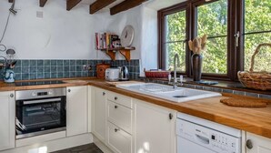 A kitchen with white cabinets, a Bosch dishwasher, a built-in oven, a window with a view of greenery, and a wooden ceiling. - Trehaverock Cottage (Port Isaac)