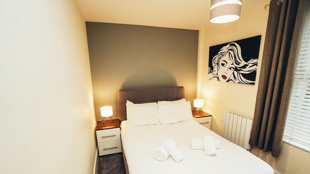 A neatly made bed with white linens and towels, a headboard, bedside lamps, a radiator, and a wall art piece.