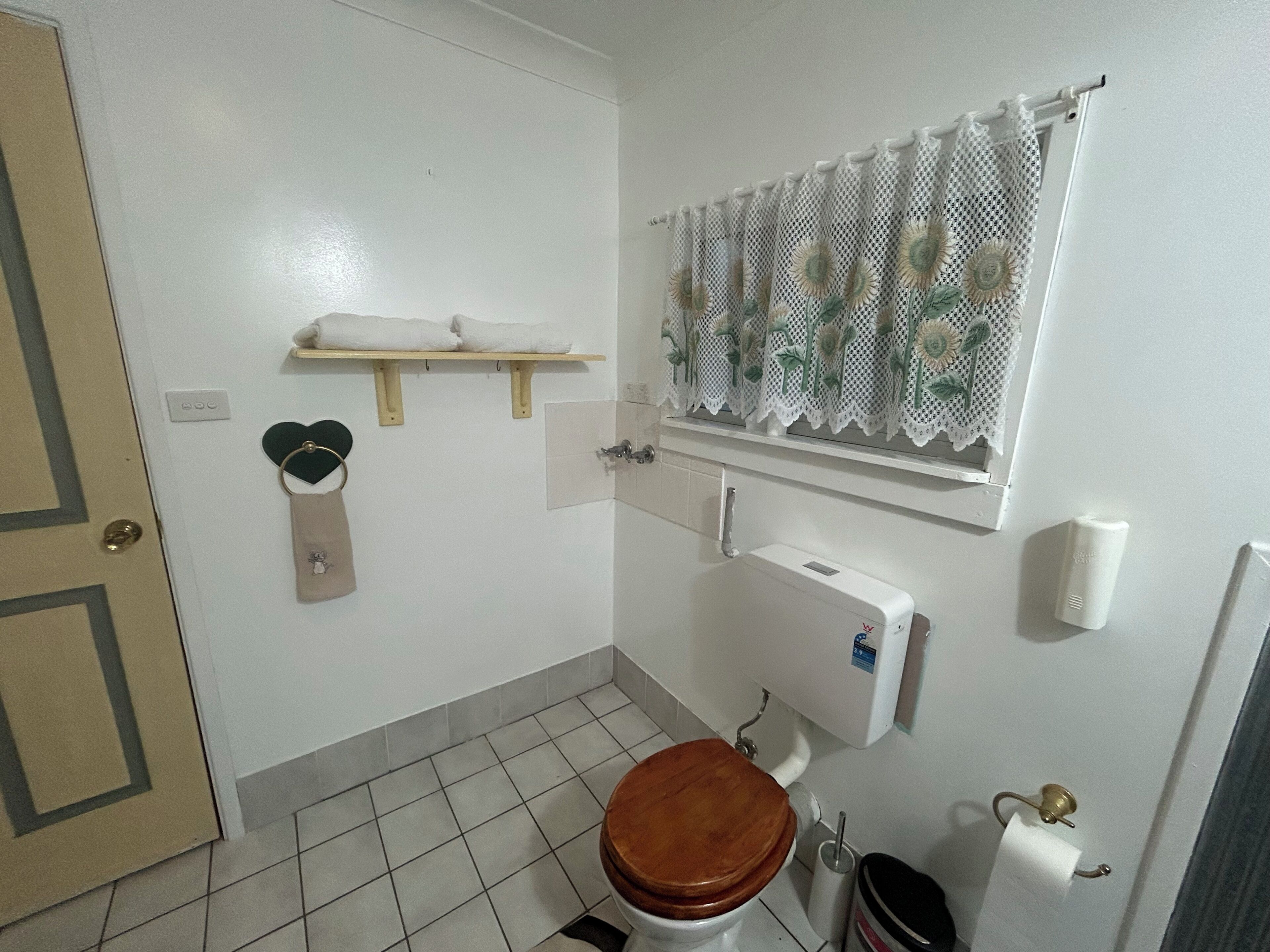 A bathroom with a toilet, a shelf with towels, a window with lace curtains, and a trash bin.