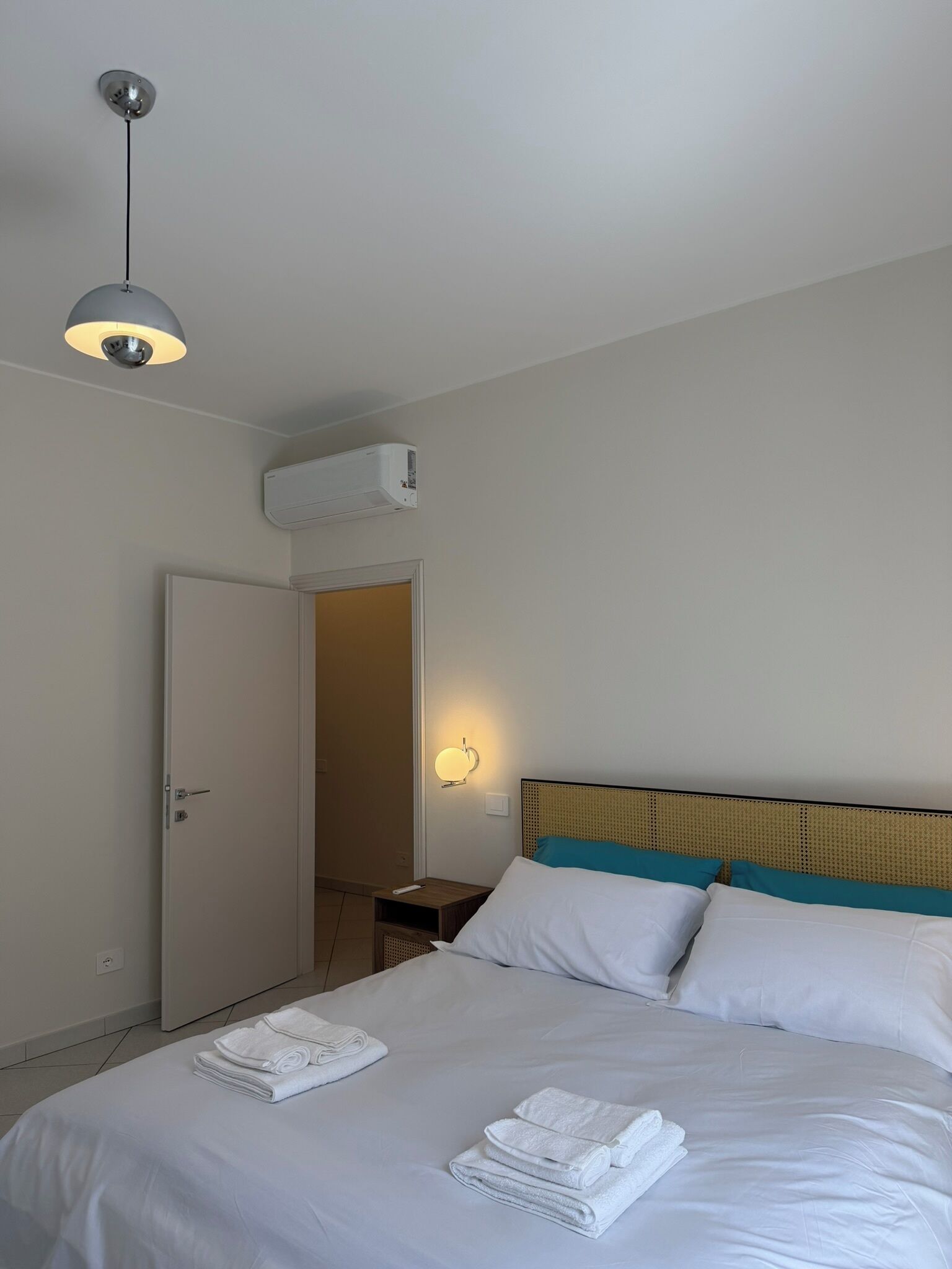 Ett hotellrum med en säng, ett sängbord, en lampa, en luftkonditioneringsanläggning och en dörr.