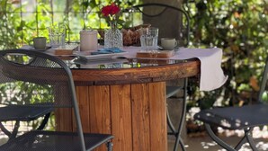 Een houten tafel met een witte tafelkleed, omringd door metalen stoelen, buiten geplaatst met een boom en begroeiing op de achtergrond.
