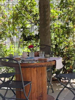 Uma mesa de madeira com toalha branca, rodeada por cadeiras de metal, instalada ao ar livre, com uma árvore e vegetação ao fundo.