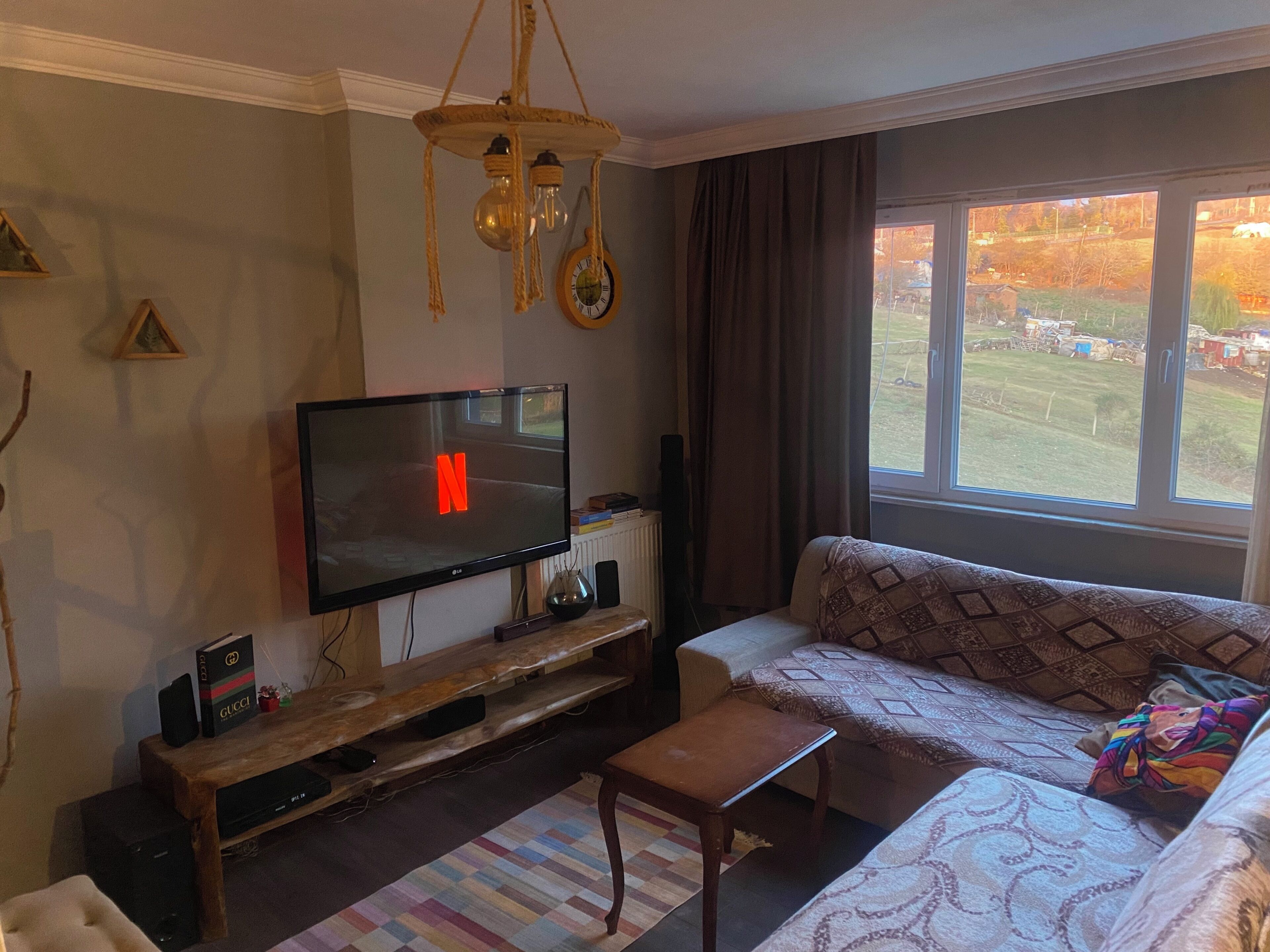 Ett vardagsrum med en platt-TV som visar Netflix-logotypen, en TV-möbel i trä, en soffa och ett fönster med utsikt över en trädgård.