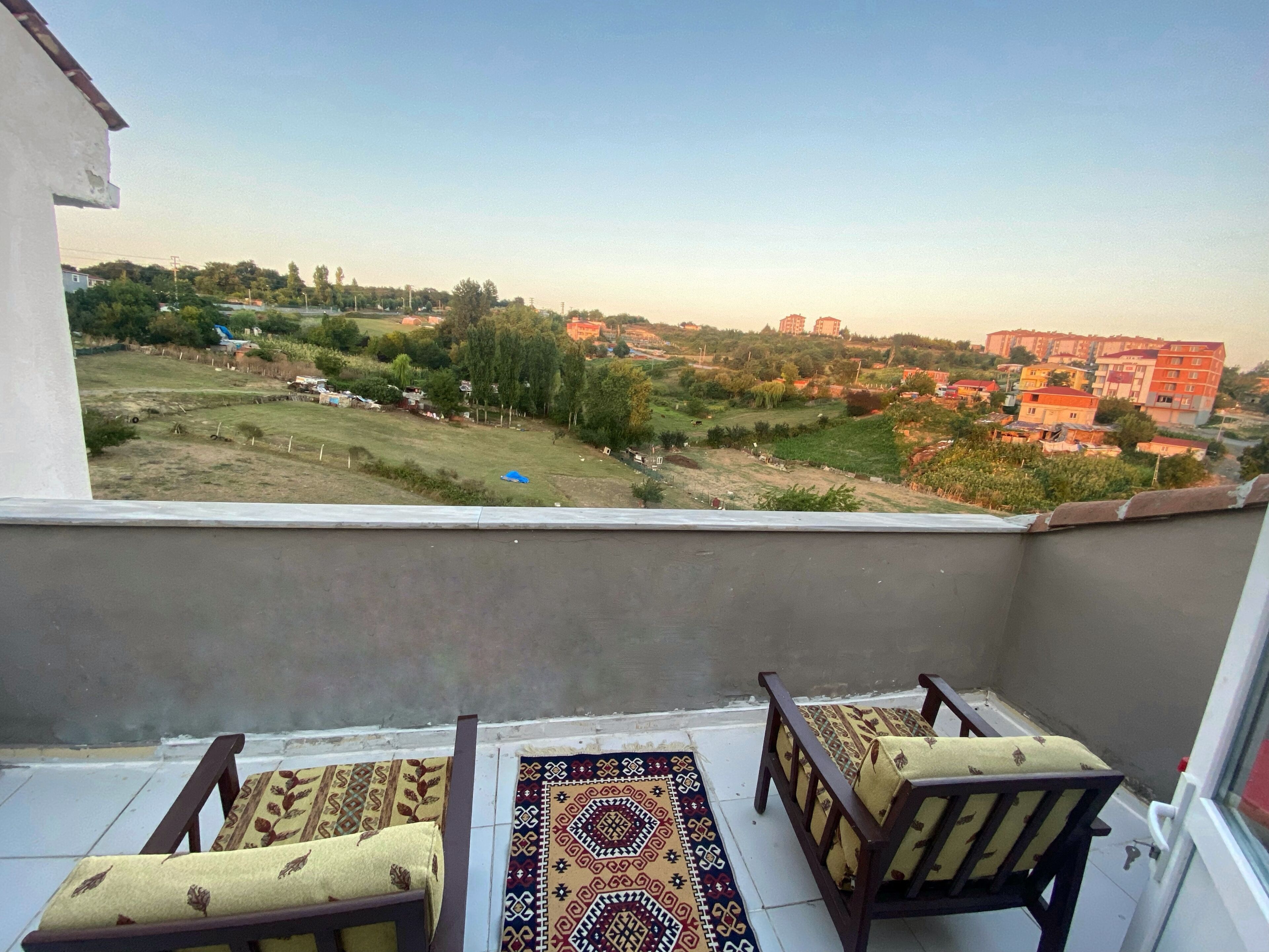 A balcony with two wooden chairs and a patterned rug, overlooking a residential area with houses and greenery.