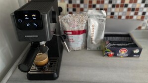 Una cafetera con pantalla digital, una taza de café y sacos de granos de café.