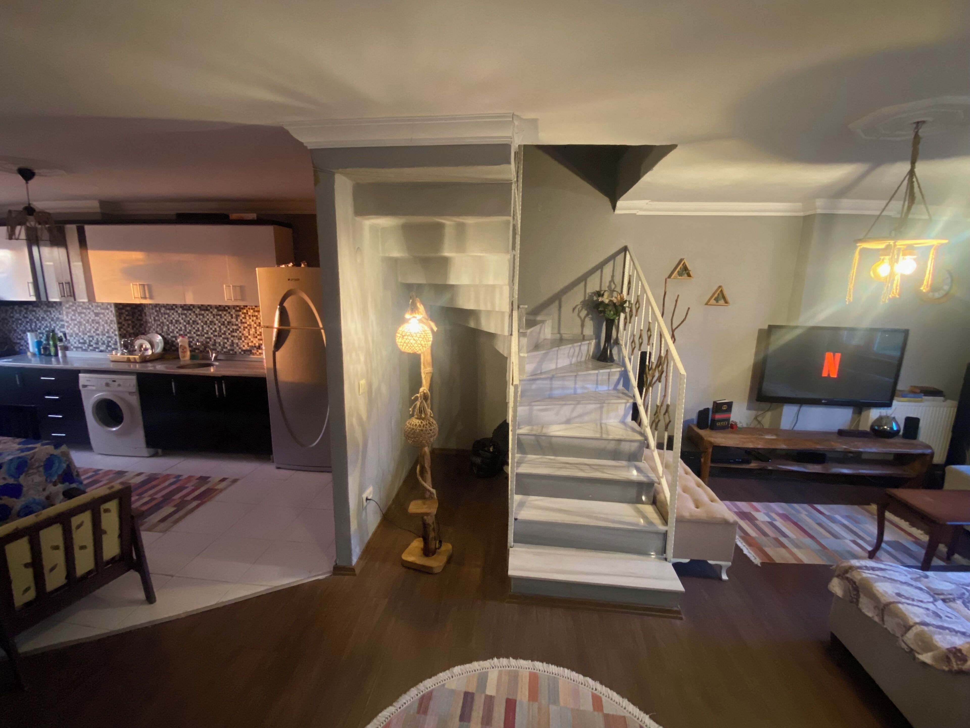 A modern living room with a staircase, a kitchen in the background, and a television displaying the Netflix logo.