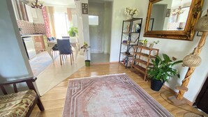 Una sala de estar con piso de madera, un tapete estampado, un sofá, una zona de comedor con sillas, una estantería y un espejo.