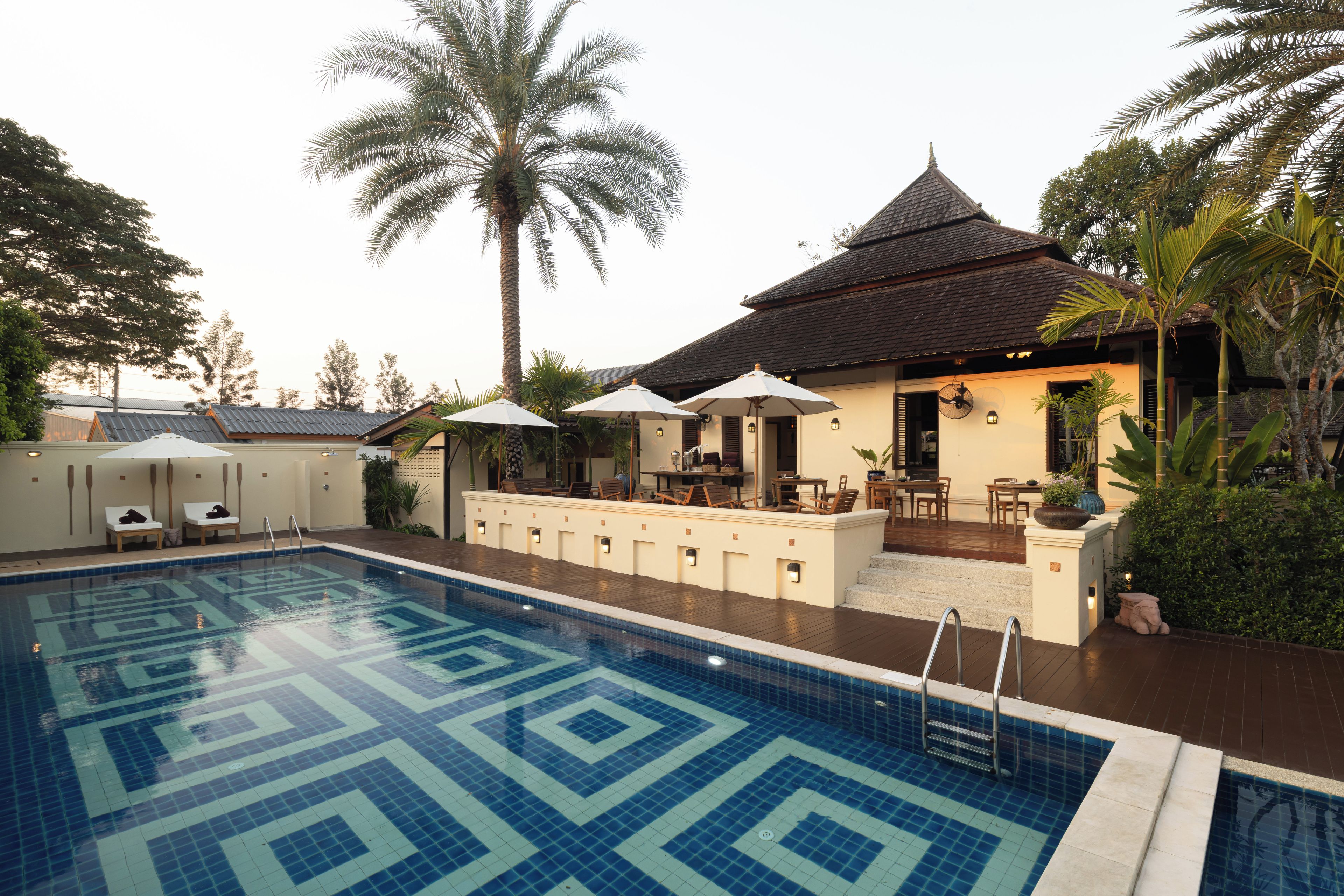 A swimming pool with a geometric tile pattern, surrounded by lounge chairs and umbrellas, with a building featuring a tiled roof and outdoor seating area.
