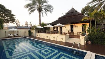 A swimming pool with a geometric tile pattern, surrounded by lounge chairs and umbrellas, with a building featuring a tiled roof and outdoor seating area.