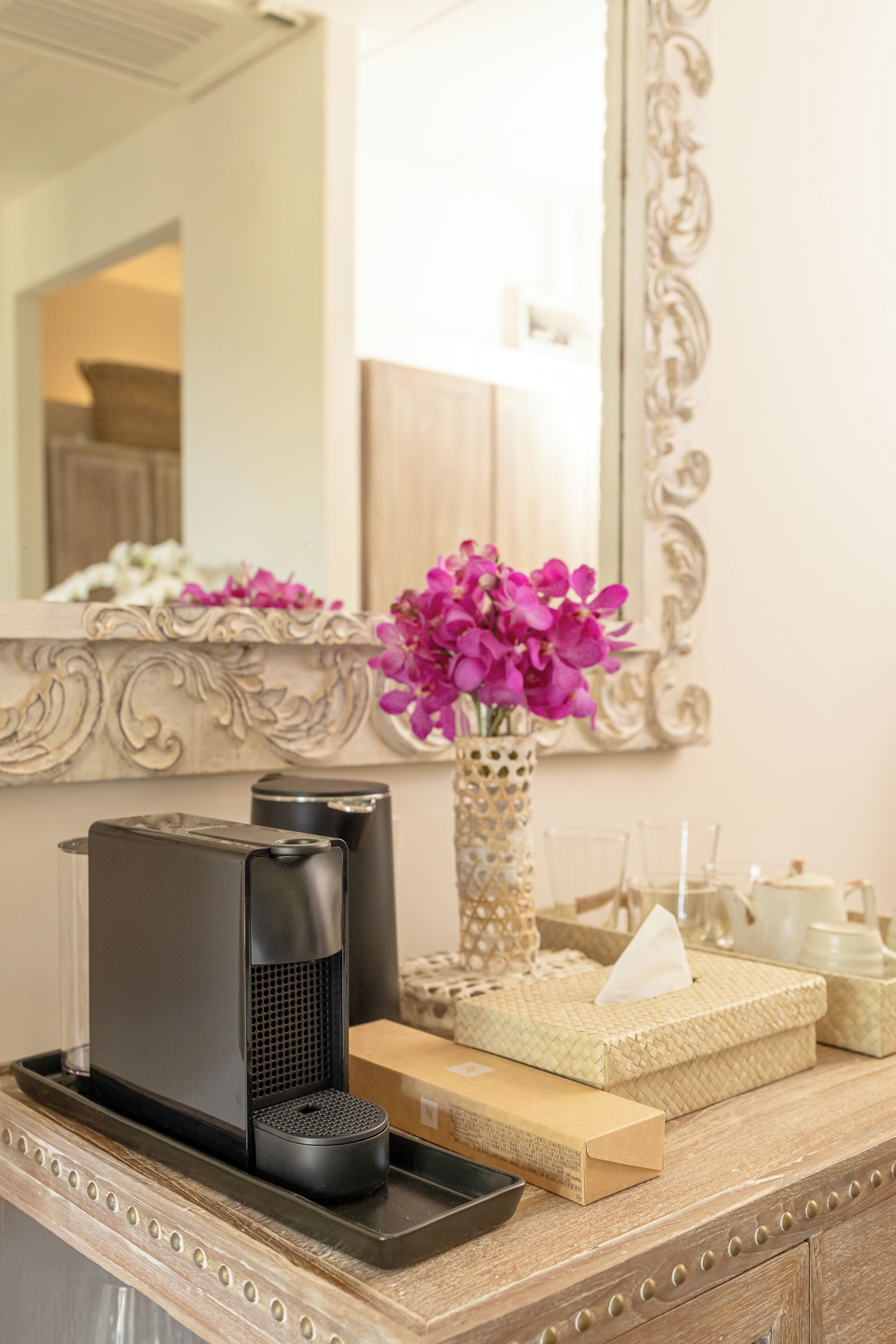 A bathroom vanity with a mirror, a vase of flowers, a coffee maker, and a tissue box.