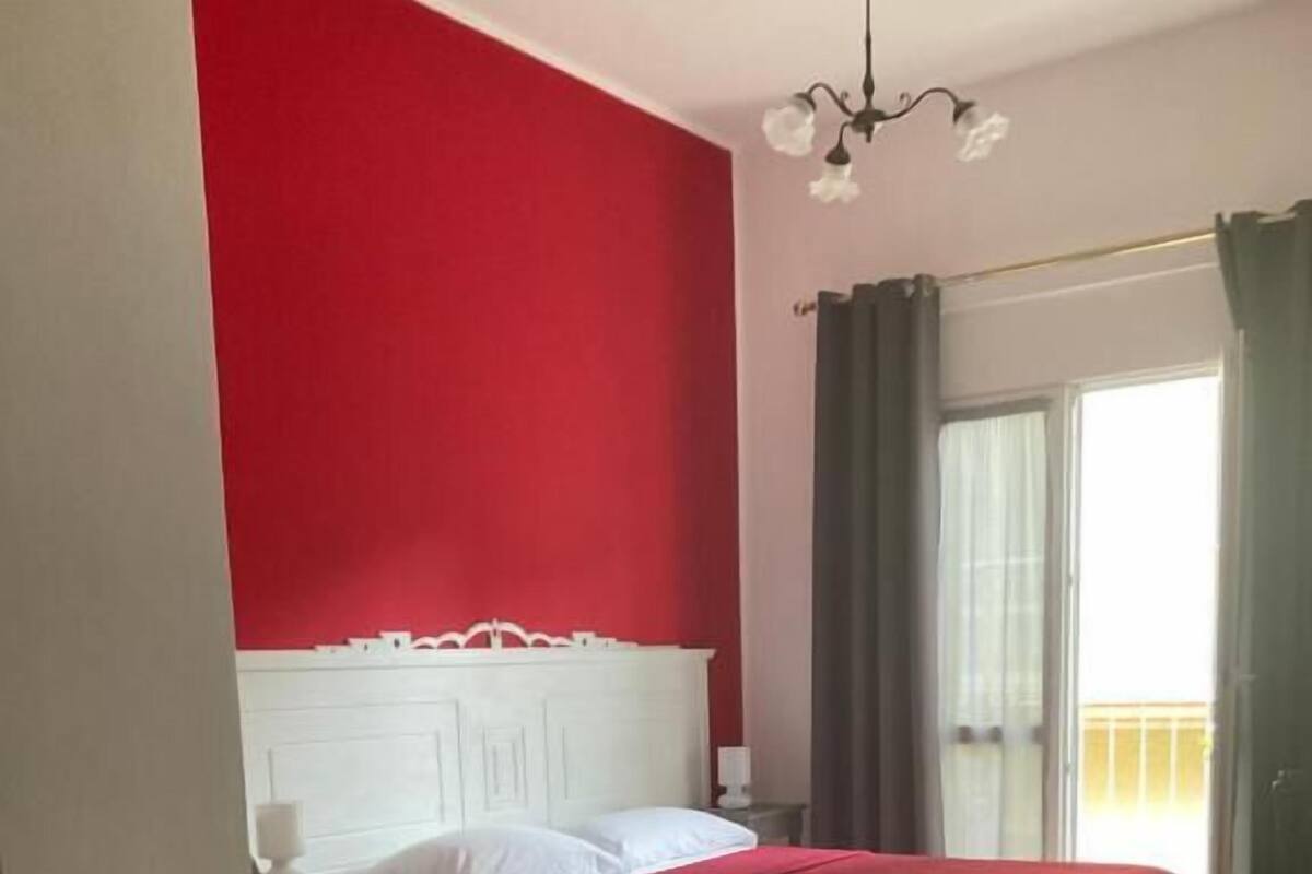 Ein Zimmer mit einem Bett, einem kleinen Tisch, einem Nachttisch, einem Stuhl und einem Fenster mit Vorhängen.