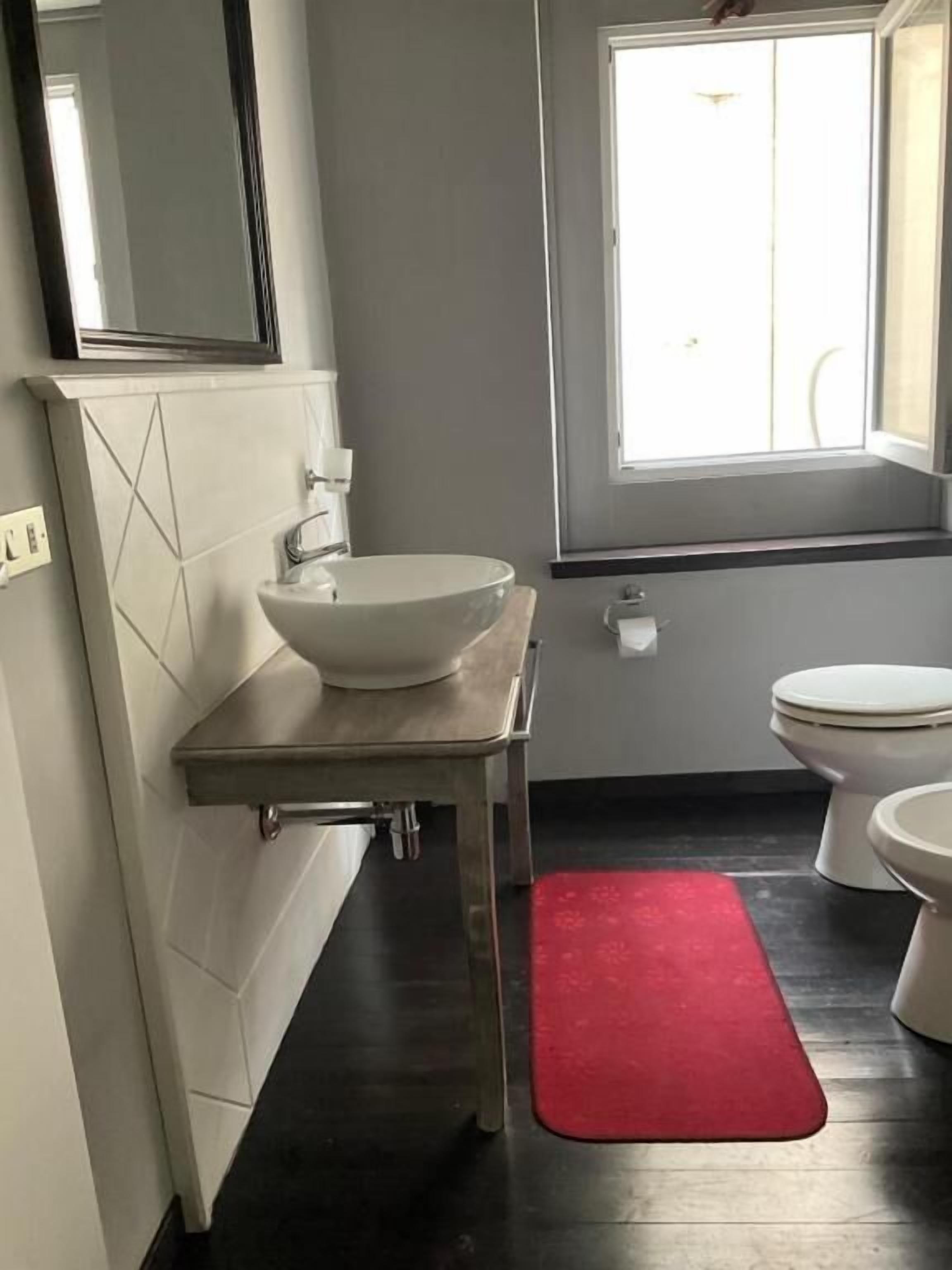 Un bany amb un lavabo blanc, un mirall, un vàter i una finestra.