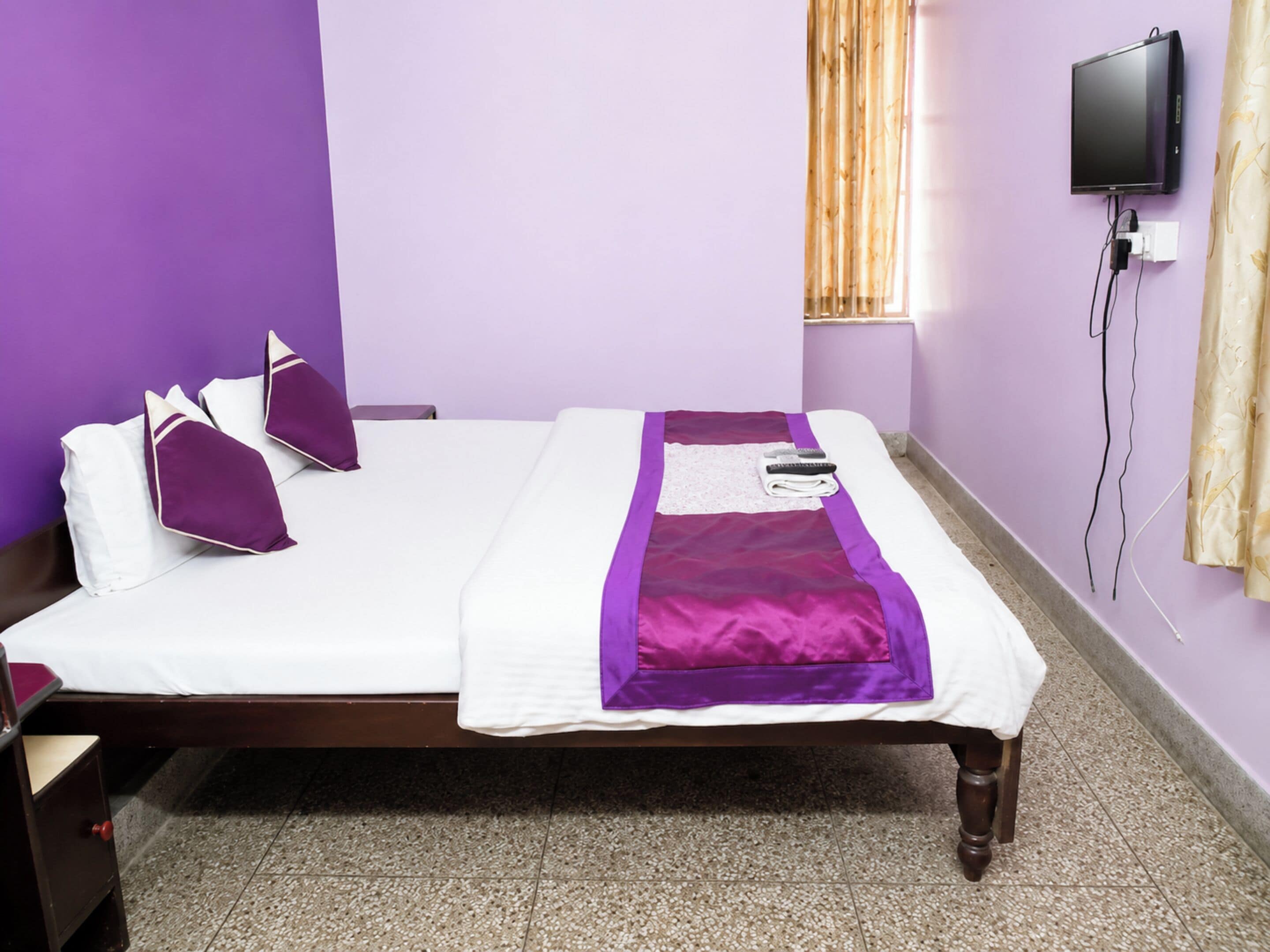 A bed with a white mattress and purple bedding, a wooden nightstand, a television mounted on the wall, and a window with curtains.