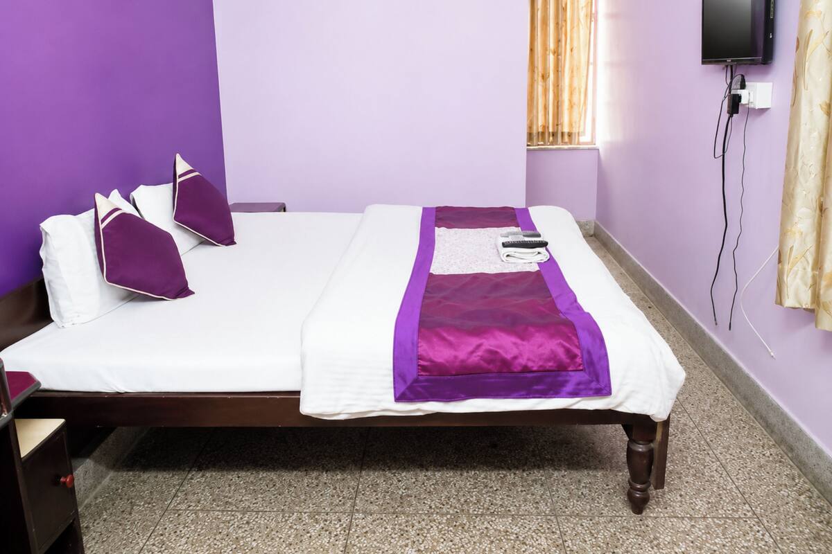 A bed with a white mattress and purple bedding, a wooden nightstand, a television mounted on the wall, and a window with curtains.