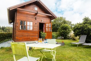 A wooden cabin with a dining table and chairs, a small garden, and a lounge chair.