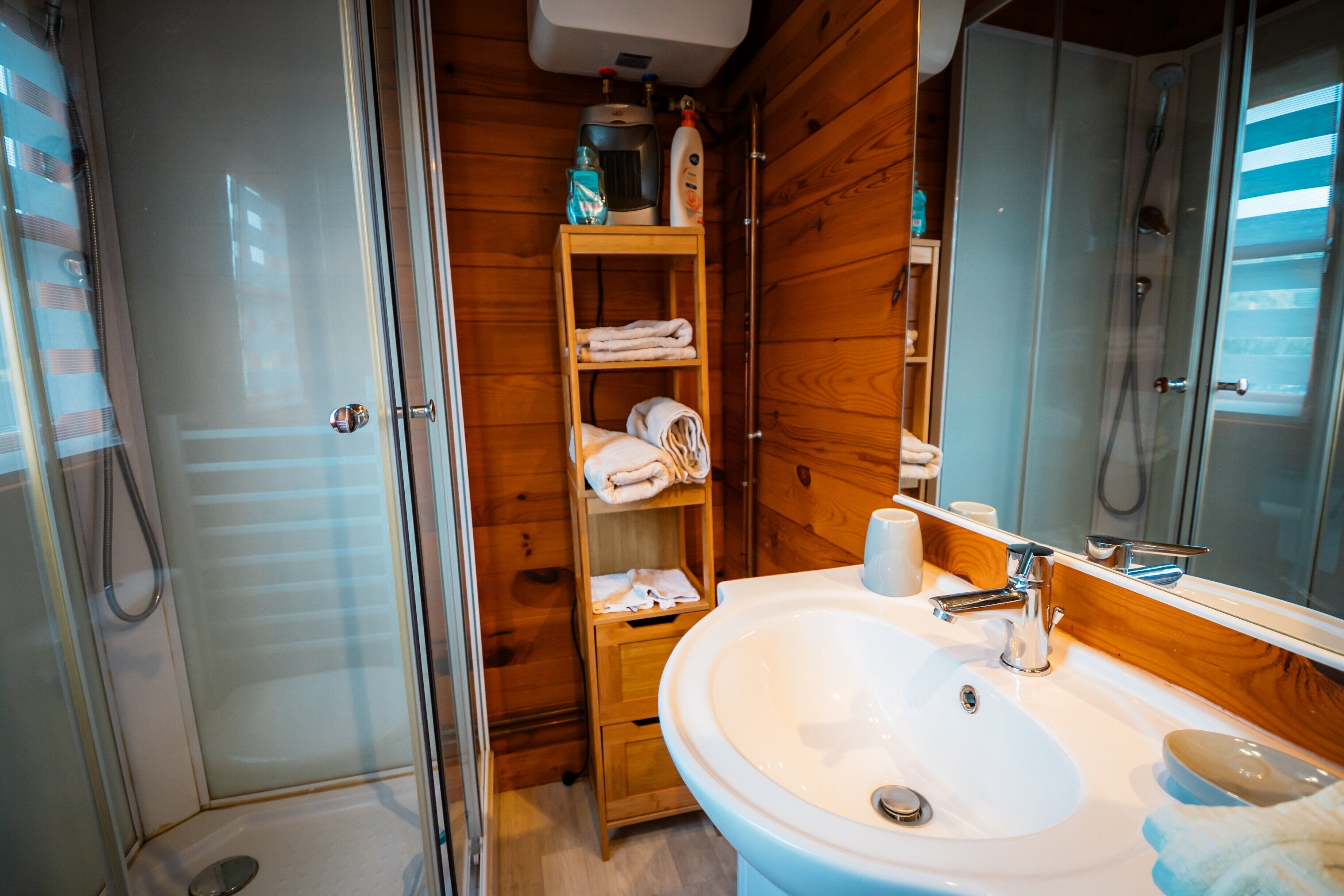 Un baño con cabina de ducha de vidrio, un estante de madera con toallas, un lavabo con espejo y un inodoro.