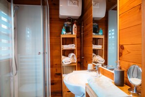 A wooden cabin bathroom with a glass shower door, a sink, a mirror, and shelves with towels and toiletries.